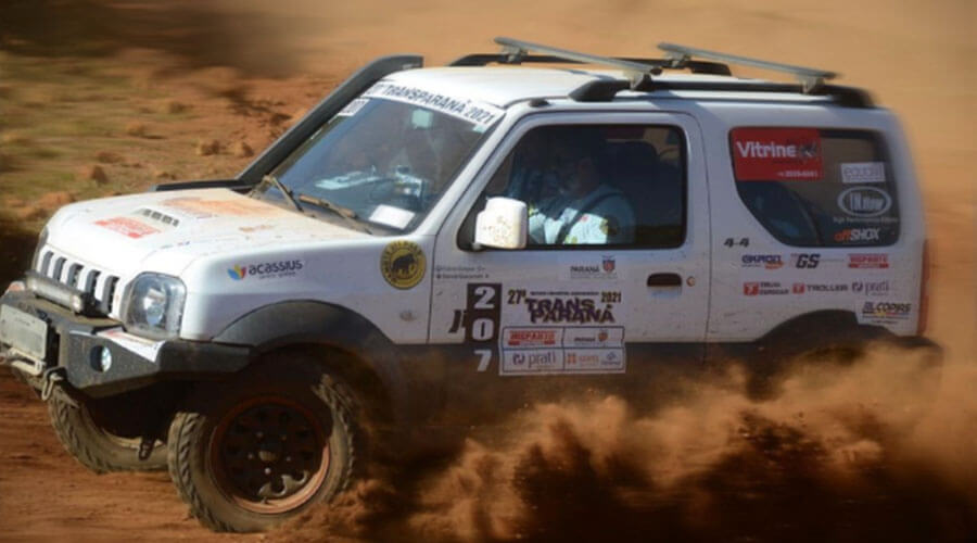 Suzuki Jinmy na cor branca com diversos adesivos colados nas laterais do veículos, carro preparado para rally, levantando poeira no deserto durante uma competição de Rally de Regularidade