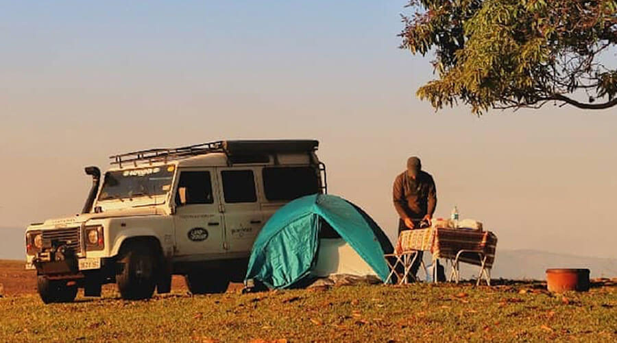 Acampamento montado em expedição de overland, carro Defender 4x4 estacional com uma barraca armada ao lado e o motorista fazendo uma refeição em churrasqueira portátel em uma paisagem árida em cima de um monte